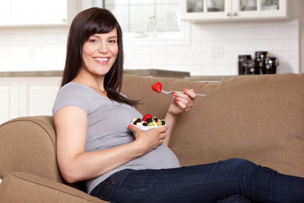 A pregnant woman eating a bowl of fruits on the couch