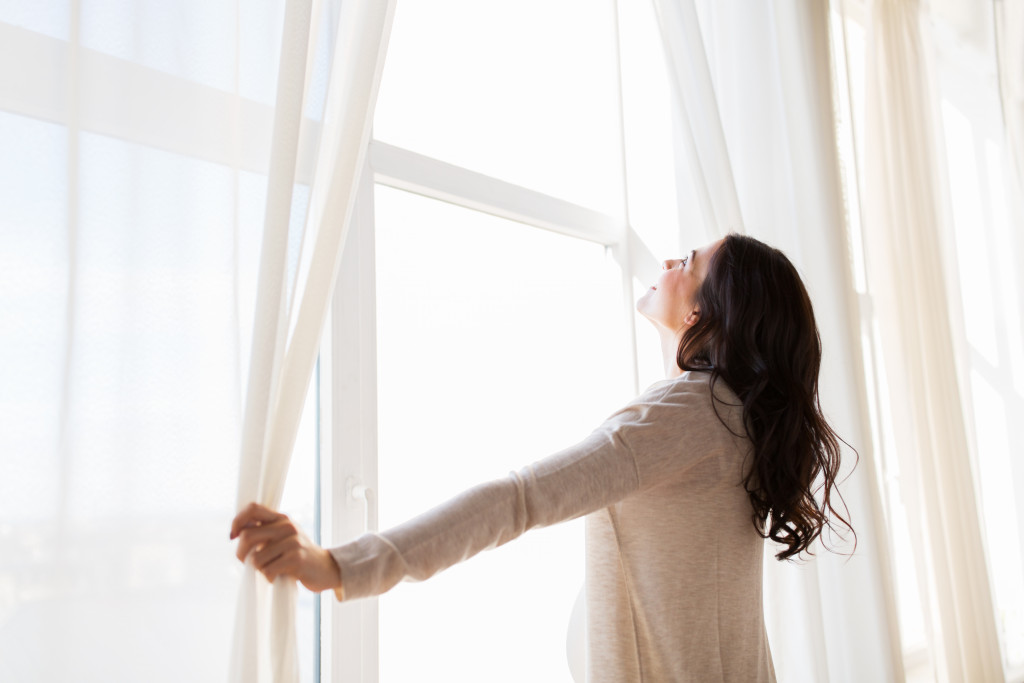 A pregnant woman opening a big curtain on a big window at home