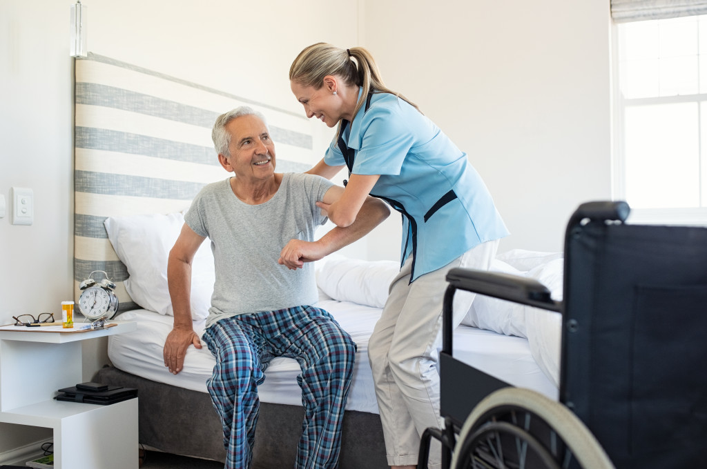 Female caregiver helping a senior man get up from his bed.